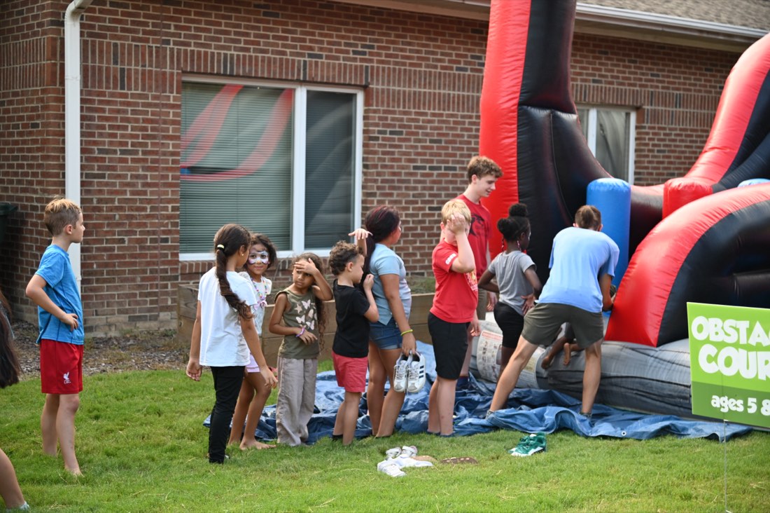 Barefoot kids lined up at an inflatable obstacle course, shoes left on the grass.