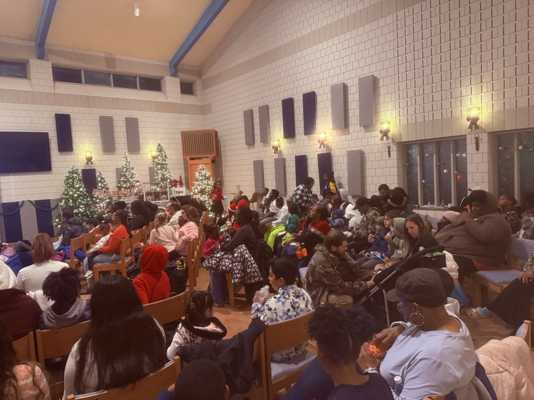 A room full of neighbors in rows of chairs, with more standing, between lit Christmas trees.